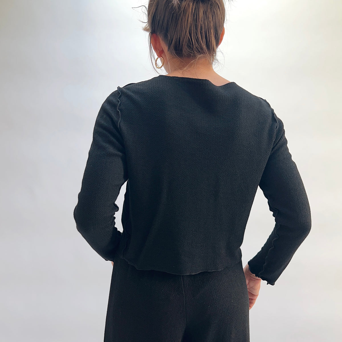 Model wearing the Cut Loose Marled Knit Cropped Cardigan in Black, back view with hands on hips, showing scalloped seam detail at shoulders, clean back panel, and cropped straight hem of black marled knit cardigan, styled with a black skirt

