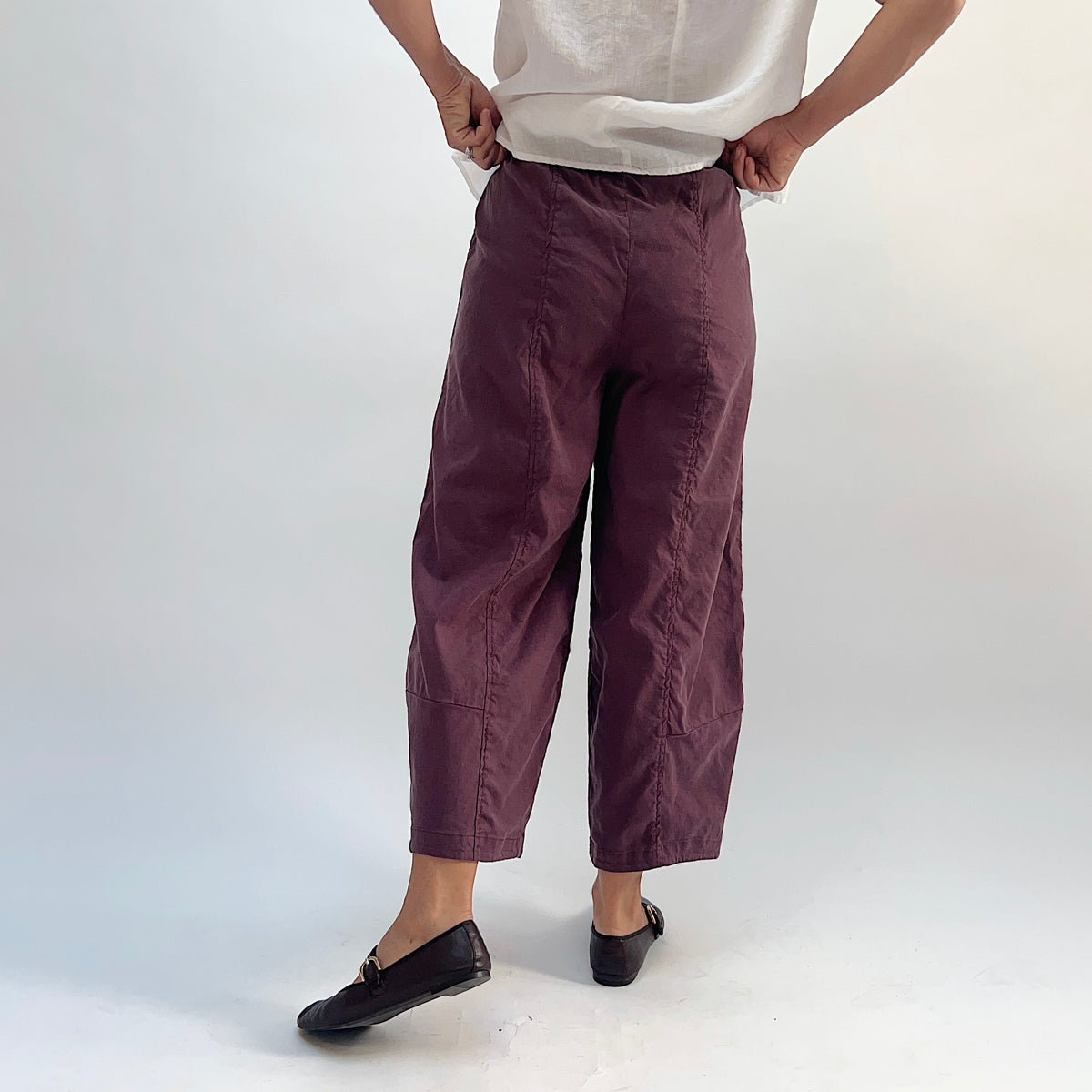 Front close-up view of russet linen-tencel lantern pants showing wide curved leg silhouette, elastic waistband, and cropped ankle length, styled with a white top and black Mary Jane flats
