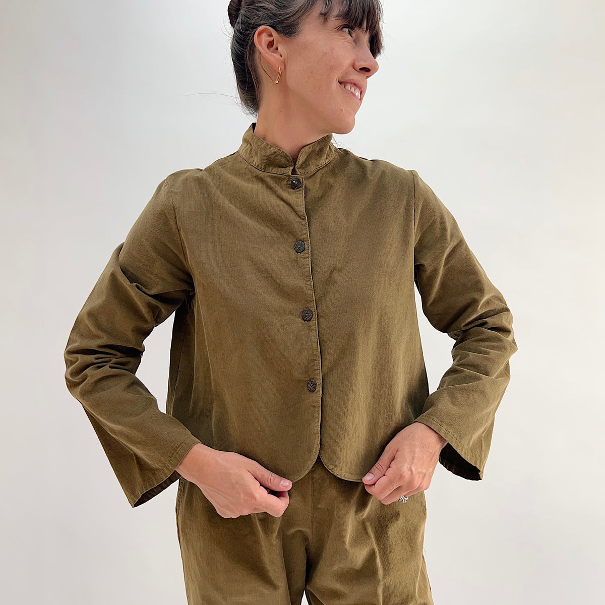 Model smiling and wearing the Bryn Walker Corduroy Standing Collar Crop Jacket in Partridge, front view showing ruffled standing collar, shell button closure, and olive pinwheel corduroy, paired with matching Pasha Pants
