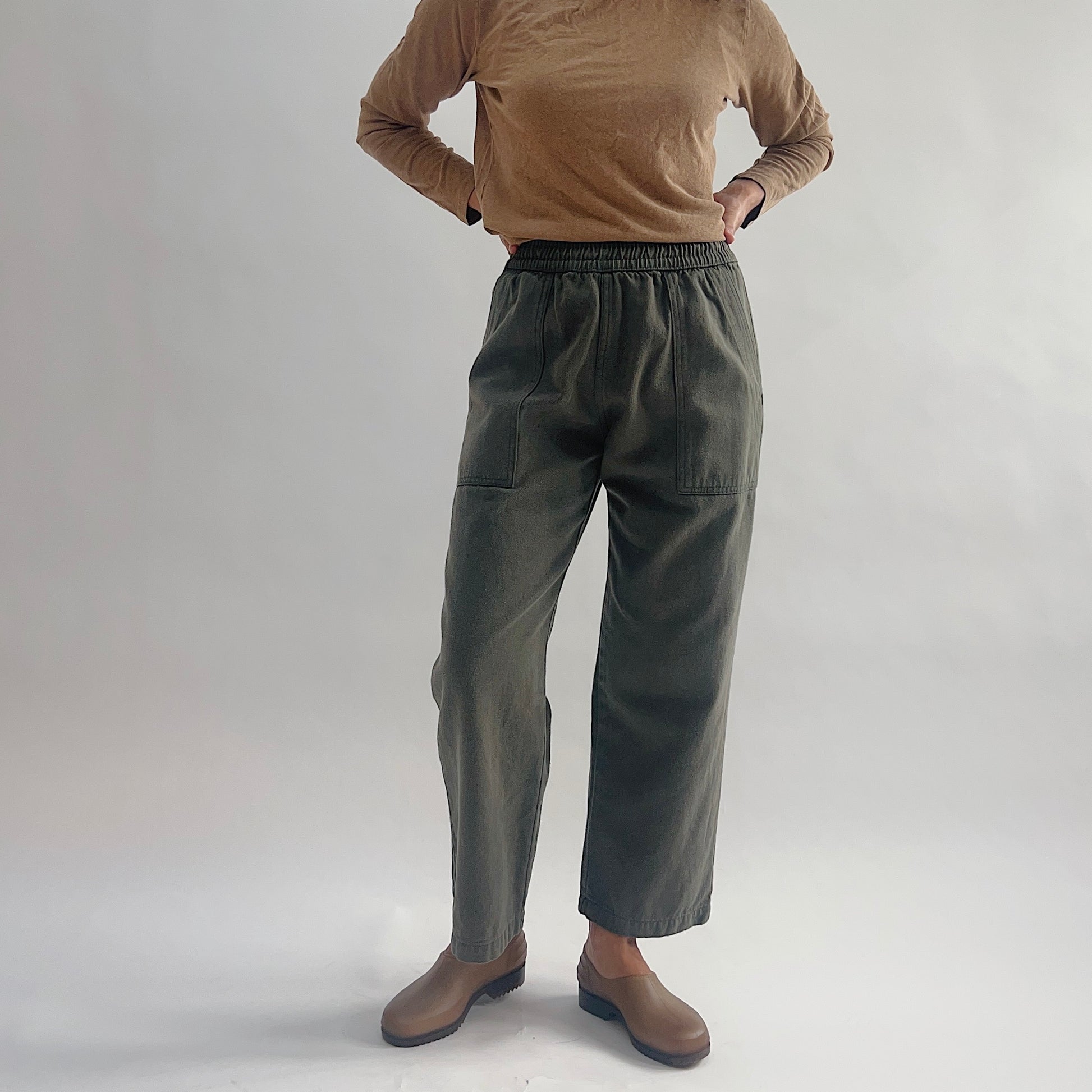 Front view of model wearing olive green high-waist wide-leg pull-on pants with two patch pockets, styled with a tan long-sleeve top and brown clogs against a white background.
