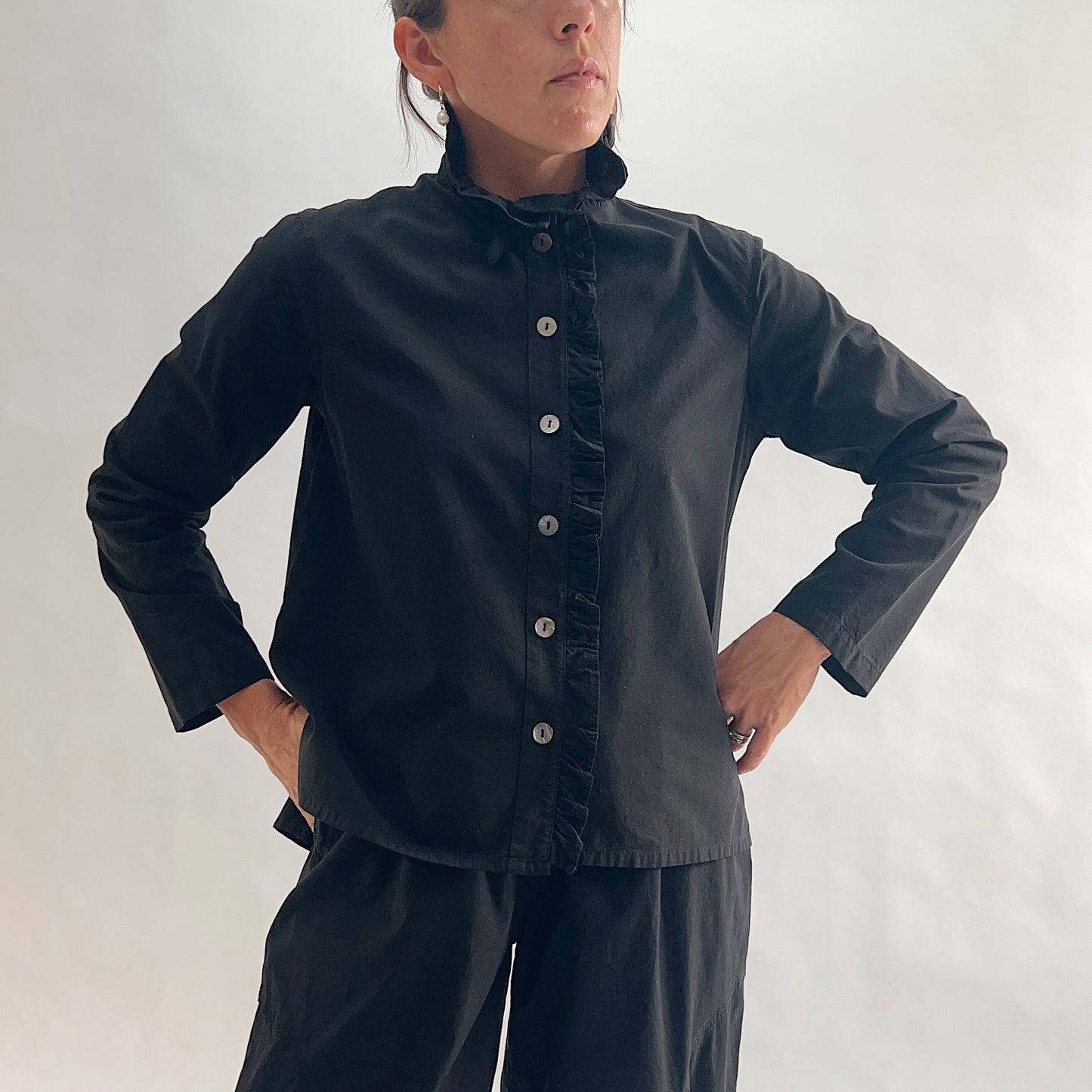 Close front view of model wearing Eleven Stitch black blossom collar shirt with hands at placket, showing ruffled blossom collar, ruffled button placket with shell buttons, long sleeves, and black color, styled with matching black wide-leg pants