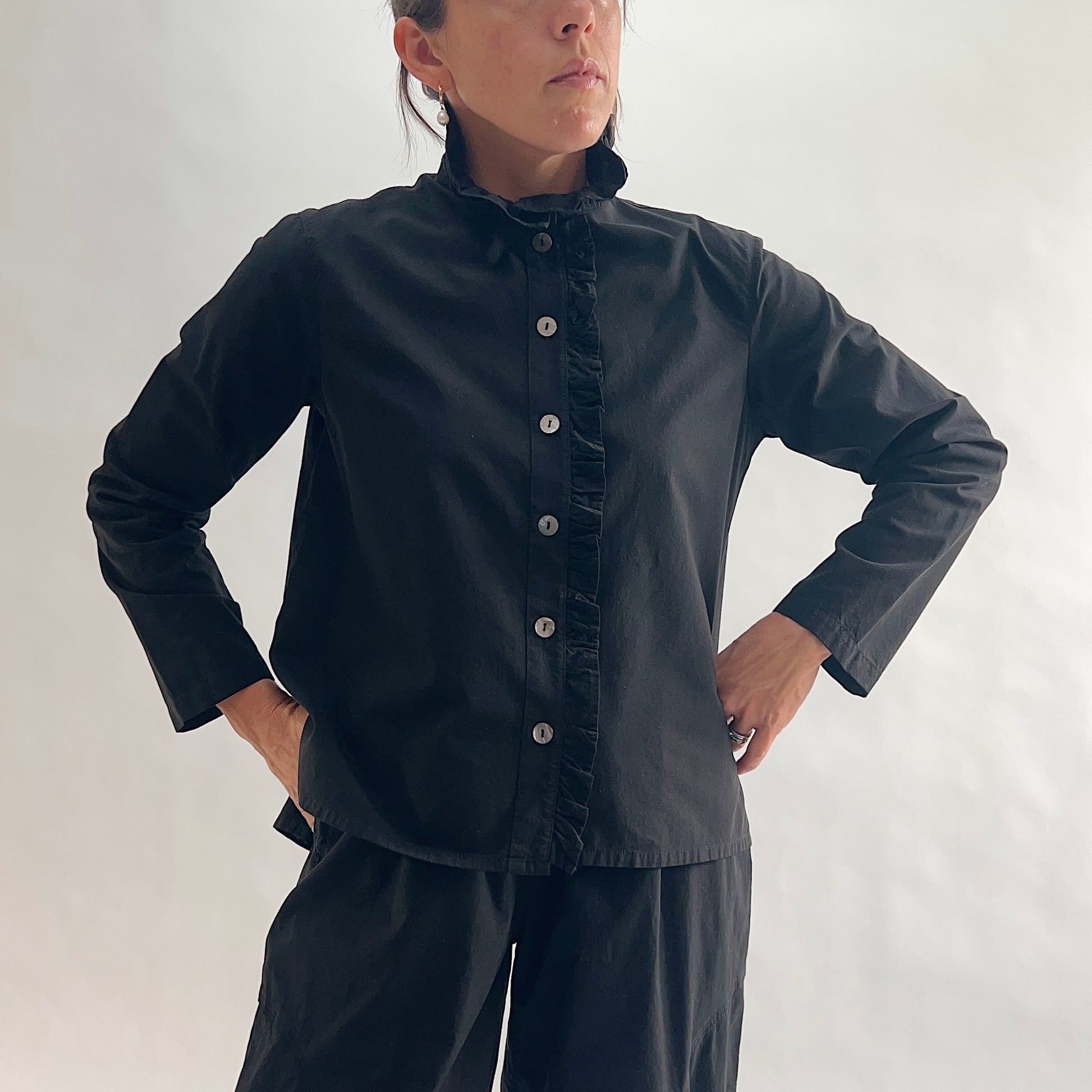 Close front view of model wearing Eleven Stitch black blossom collar shirt with hands at placket, showing ruffled blossom collar, ruffled button placket with shell buttons, long sleeves, and black color, styled with matching black wide-leg pants