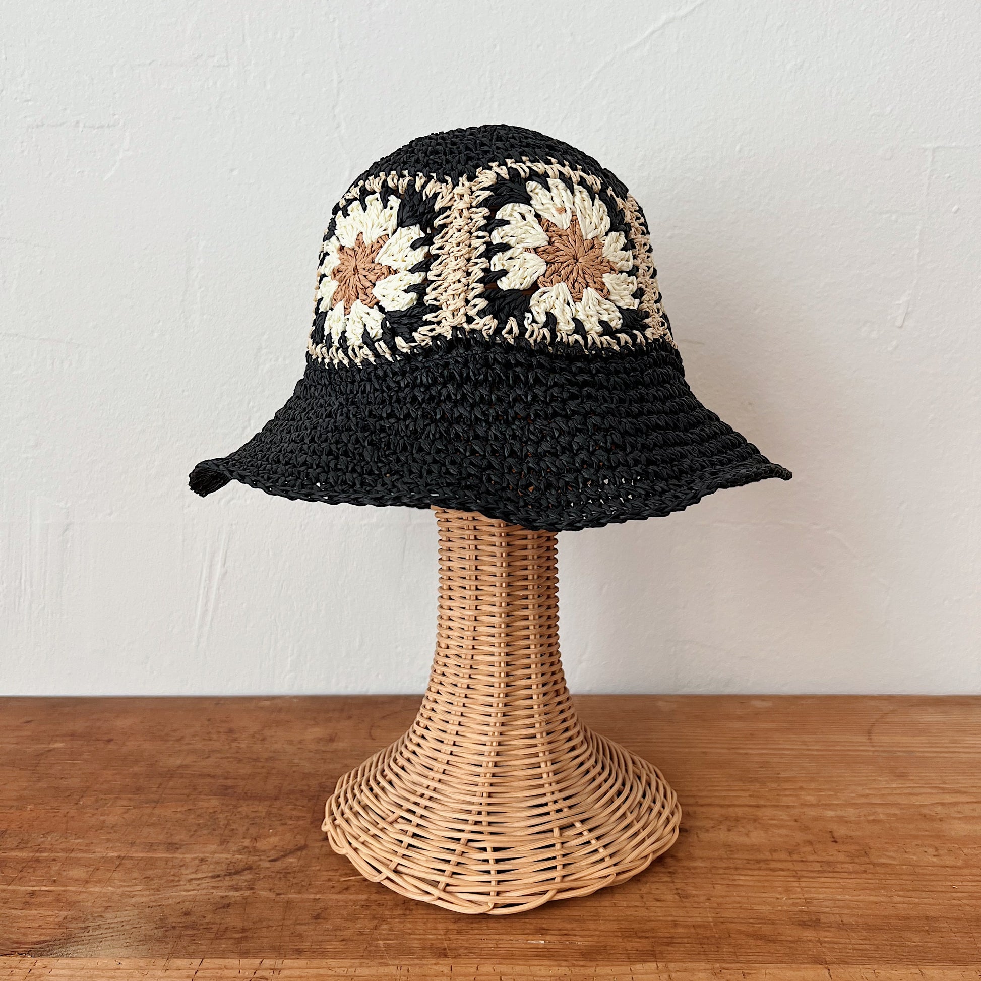 Black paper crochet bucket hat with cream and tan floral granny square band, shown on a wicker hat stand — front view