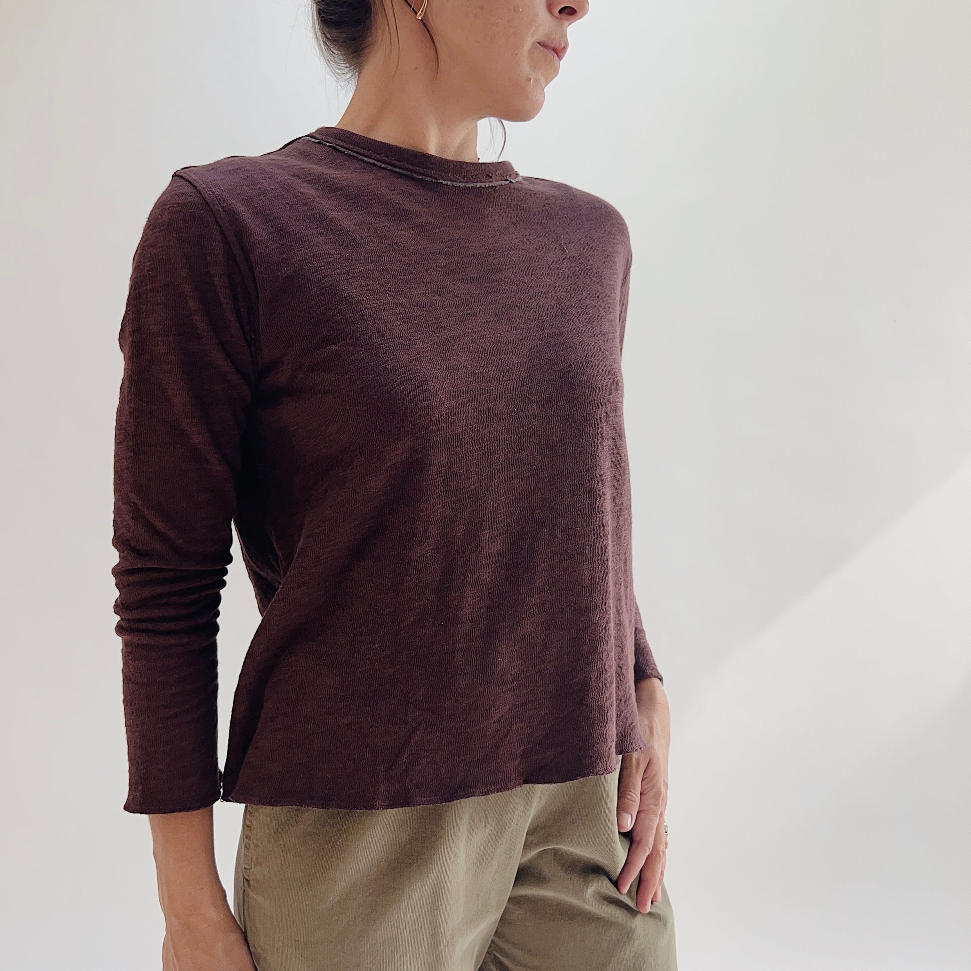 Model wearing the Bryn Walker Double Cotton Tee in Chicory, angled front view showing raw-edge crew neckline, double-layer texture, relaxed silhouette, and slightly cropped long sleeves in deep chicory burgundy, styled with olive pants
