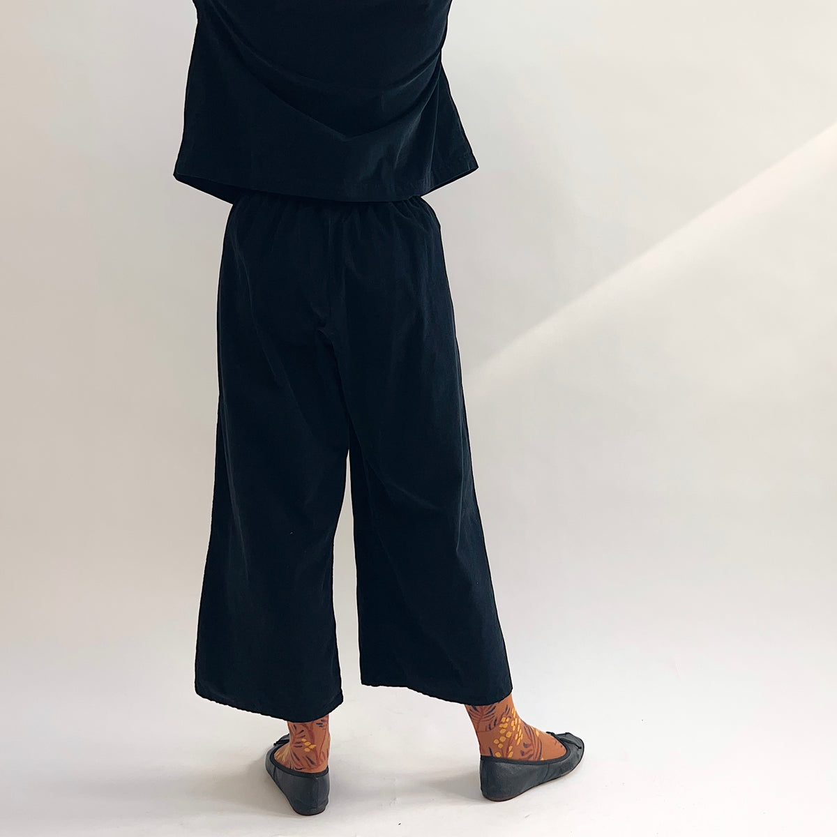 Back view of model wearing the Bryn Walker Corduroy Flood Pants in Black, showing elastic waistband, wide-leg silhouette, flood-length hem, and black pinwheel corduroy, paired with matching jacket, printed socks, and black ballet flats
