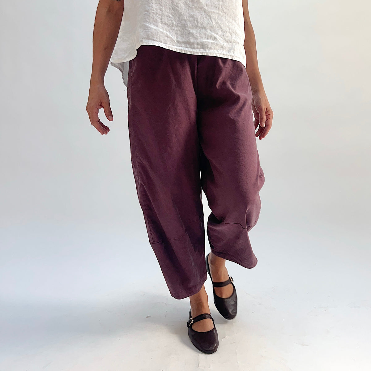 Front close-up view of russet linen-tencel lantern pants showing wide curved leg silhouette, elastic waistband, and cropped ankle length, styled with a white top and black Mary Jane flats
