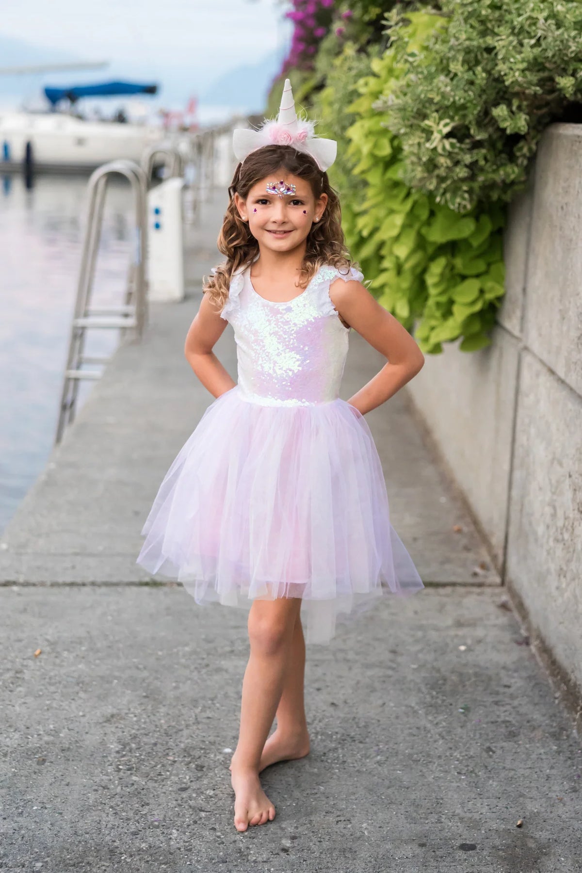 Child wearing the Great Pretenders Dreamy Unicorn Dress and Headband, full-length view showing the iridescent sequin bodice and layered pink tulle skirt, with the unicorn horn headband, outdoors

