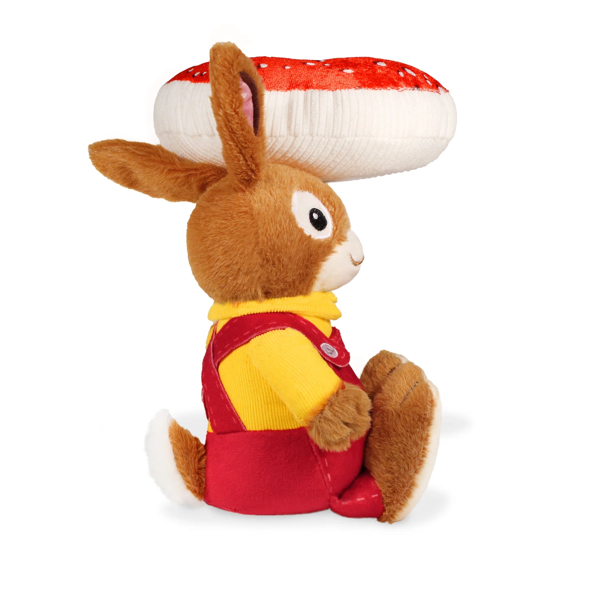 Side view of the Nicholas bunny plush toy wearing red overalls and a yellow shirt, with the red-and-white spotted toadstool balanced above his head, against a white background.
