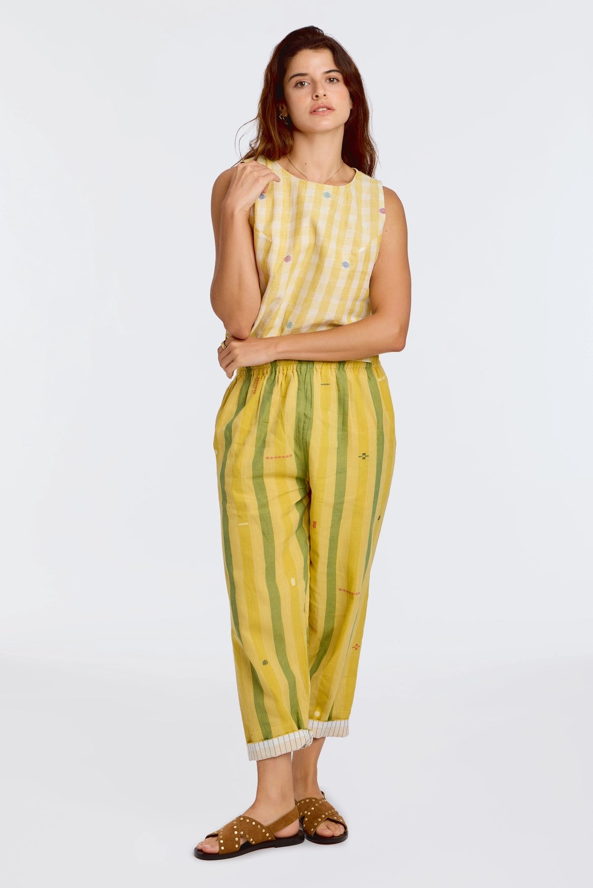 Model wearing DVAA Celeste Pant in Goldenrod, full front view against white studio backdrop showing goldenrod yellow and sage green wide vertical stripe handwoven cotton with scattered ikat motifs, elasticated drawstring waist, cropped tapered leg with contrast white pinstripe cuff turn-up, arms crossed, styled with matching yellow handwoven sleeveless crop top and tan gold-studded slide sandals

