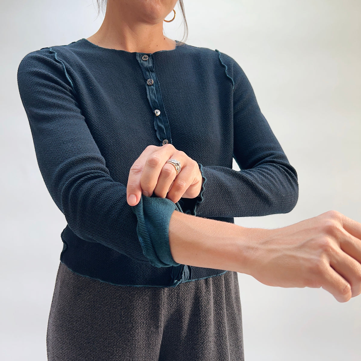 Close-up of model wearing the Cut Loose Marled Knit Cropped Cardigan in Etan, rolling up sleeve to show contrasting teal inner cuff lining and scalloped lettuce-edge cuff detail, with iridescent shell button placket visible
