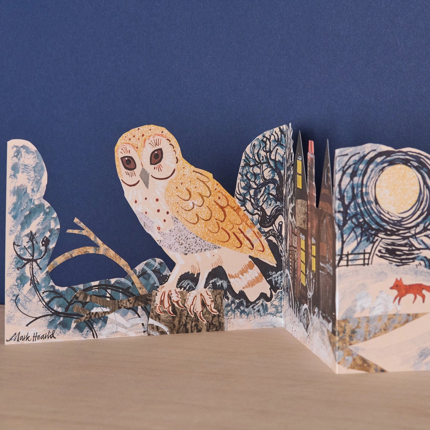Close-up detail of the Three Fold Silent Flight concertina card showing a golden barn owl perched on a branch in a snowy winter landscape, with a lit cottage and a red fox in the distance, signed by Mark Hearld