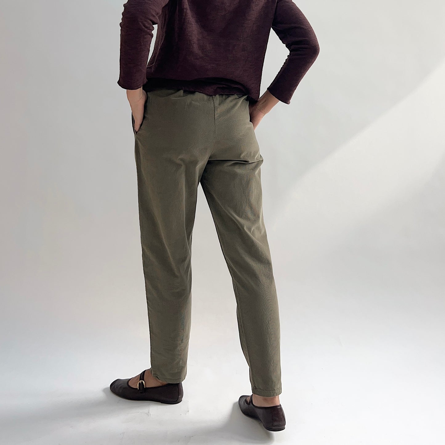 Back view of model wearing the Bryn Walker Corduroy Boslow Pants in Jasper, showing elastic waistband, relaxed tapered leg, ankle length, and olive jasper pinwheel corduroy, styled with burgundy Double Cotton Tee and brown Mary Jane flats
