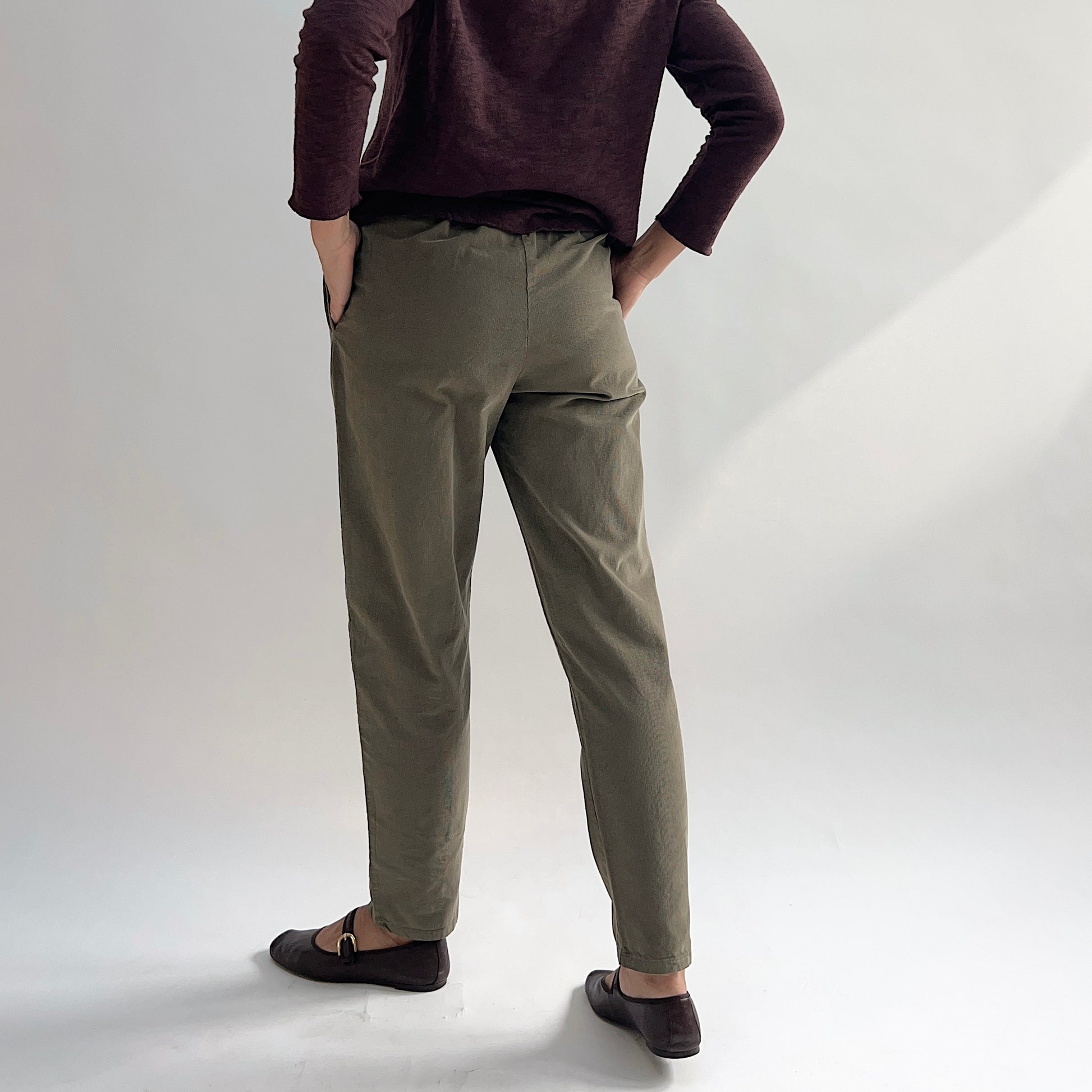 Back view of model wearing the Bryn Walker Corduroy Boslow Pants in Jasper, showing elastic waistband, relaxed tapered leg, ankle length, and olive jasper pinwheel corduroy, styled with burgundy Double Cotton Tee and brown Mary Jane flats
