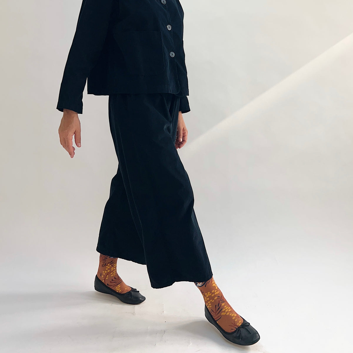 Model in motion wearing the Bryn Walker Corduroy Flood Pants in Black, side view highlighting wide-leg drape, flood-length hem, and black pinwheel corduroy texture, paired with matching Standing Collar Crop Jacket, printed socks, and black ballet flats
