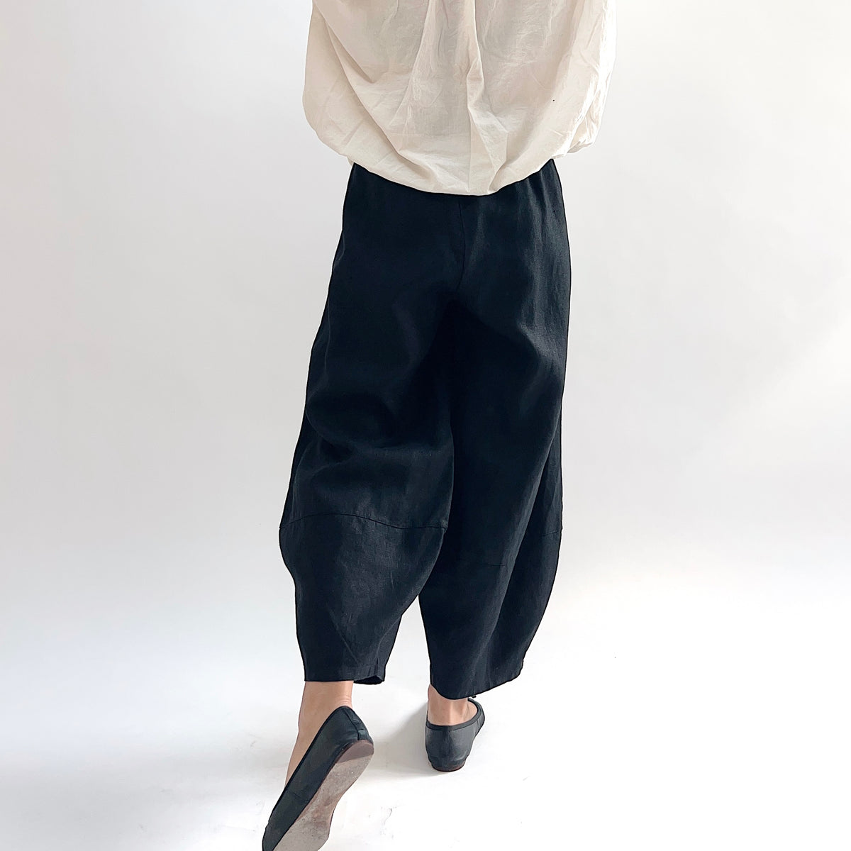 Back view of model wearing the Bryn Walker Heavy Linen Oliver Pant in Black, showing elastic waistband, barrel-leg drape, seam panel detail at knee, and tapered ankle in black heavy linen, styled with cream linen top and black slip-on flats
