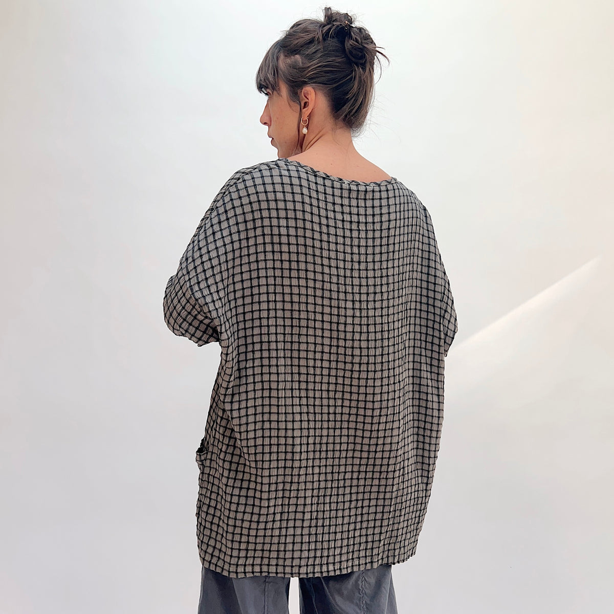 Front smiling view of squirrel grid-check crinkle-texture one-size pocket tunic with hands in both front patch pockets, showing scoop neckline with contrast stitched trim, long sleeves, and relaxed boxy fit, paired with grey wide-leg pants

