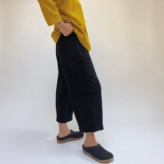 Side lower-body view of a woman wearing the Bryn Walker Pasha Fleece Pant in Black, showing the elastic waistband, large thigh patch pocket, wide straight leg silhouette, and folded ankle hem in black organic cotton and bamboo fleece, styled with a mustard yellow sweatshirt and dark slide clogs against a white background
