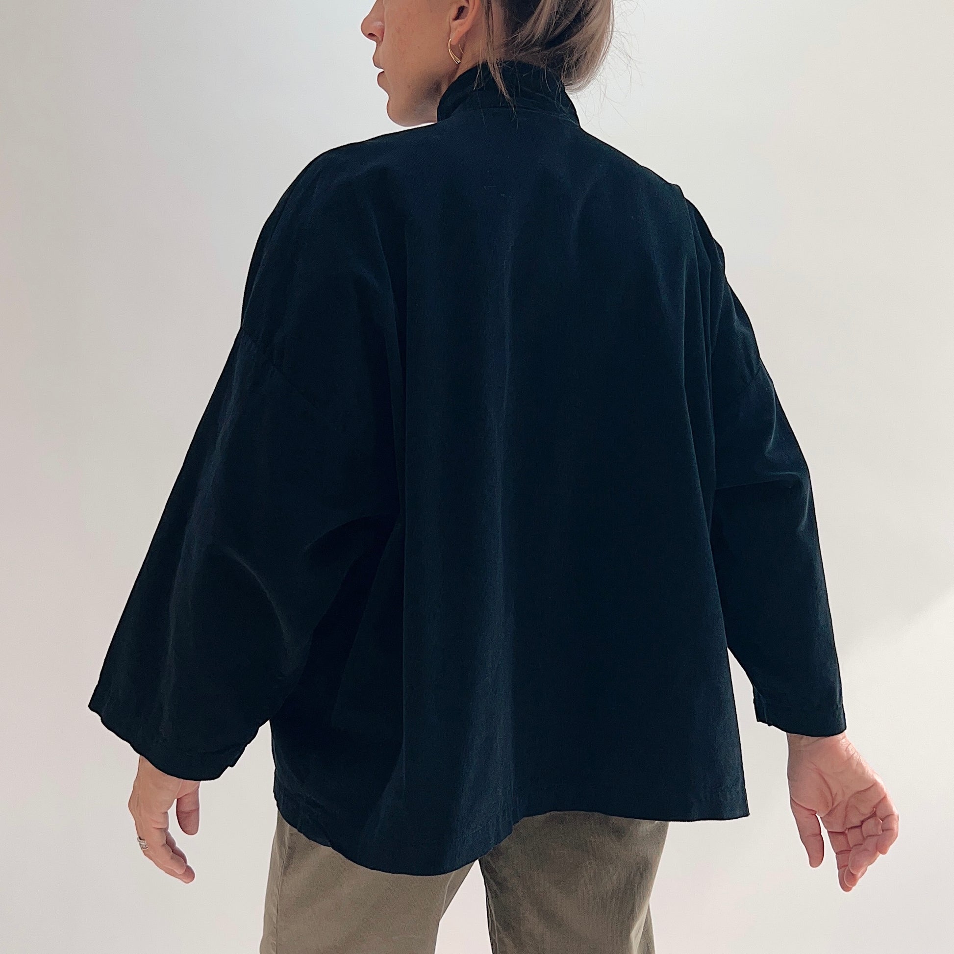 Back view of model wearing the Bryn Walker Corduroy Danuta Jacket in Black, showing standing collar, wide dolman sleeves, curved longer hem, and black pinwheel corduroy drape, styled with olive pants
