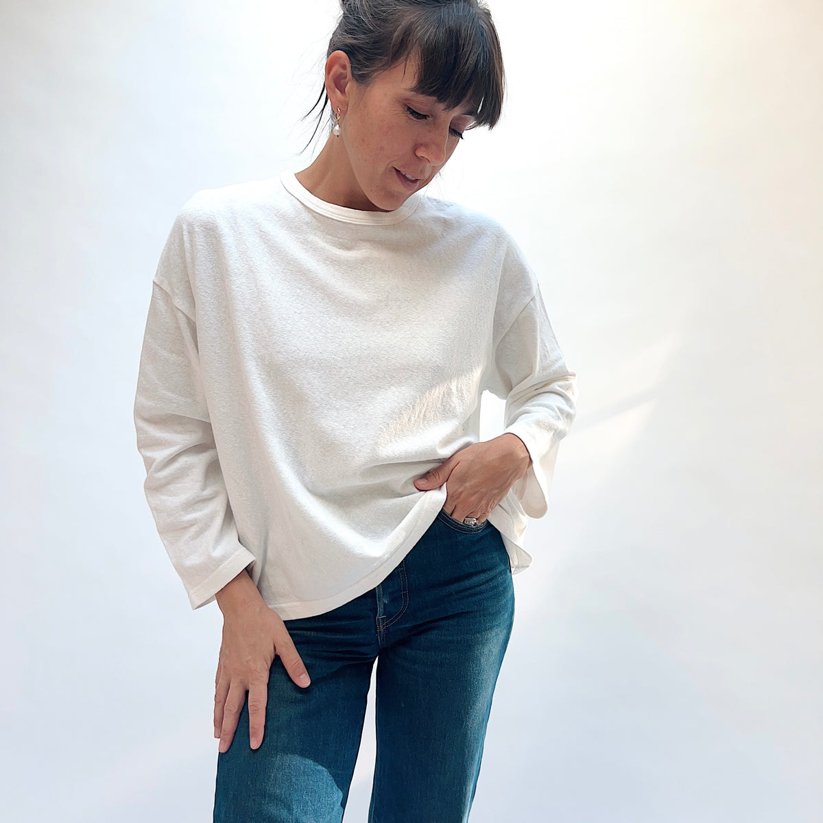Model looking down while wearing the white cotton knit pullover with blue jeans, holding one side of the hem.
