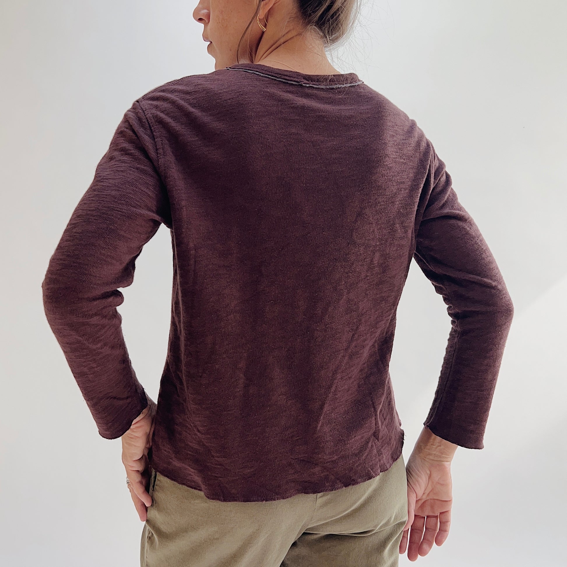 Back view of model wearing the Bryn Walker Double Cotton Tee in Chicory, showing raw-edge neckline, double-layer cotton texture, relaxed silhouette, and long sleeve length in deep chicory burgundy, styled with olive pants
