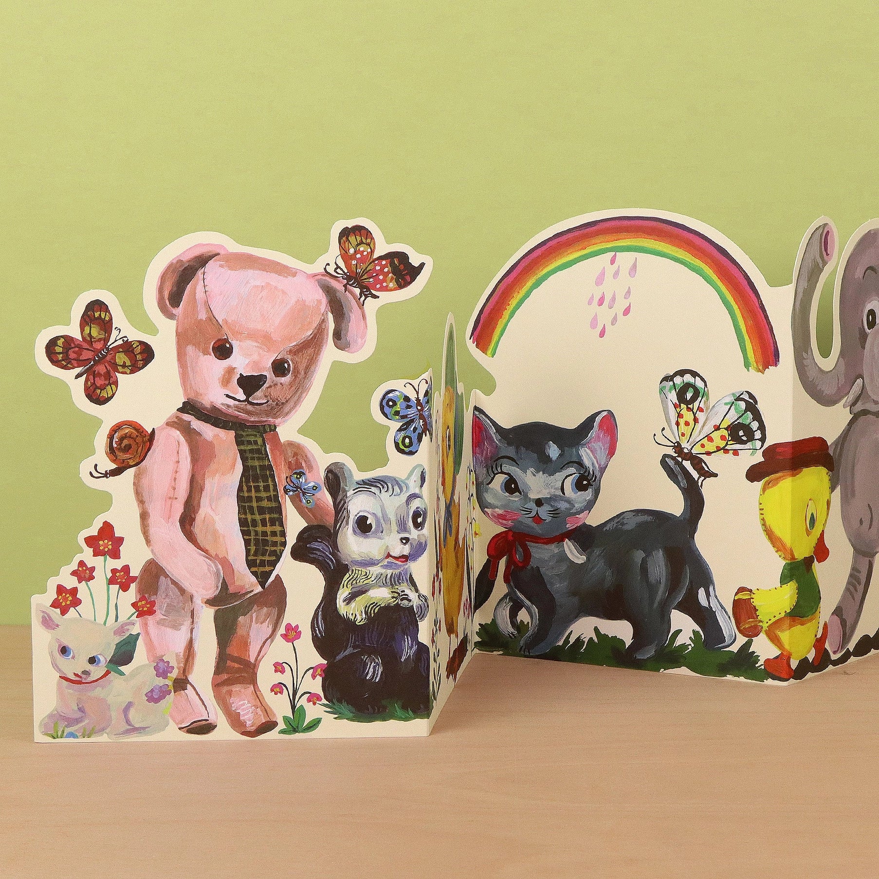 Plushie Party die-cut concertina card by Nathalie Lété, fully extended to show a parade of vintage-style plush animals including a pink bear, skunk, duck, black cat, chick, and elephant with butterflies and a rainbow