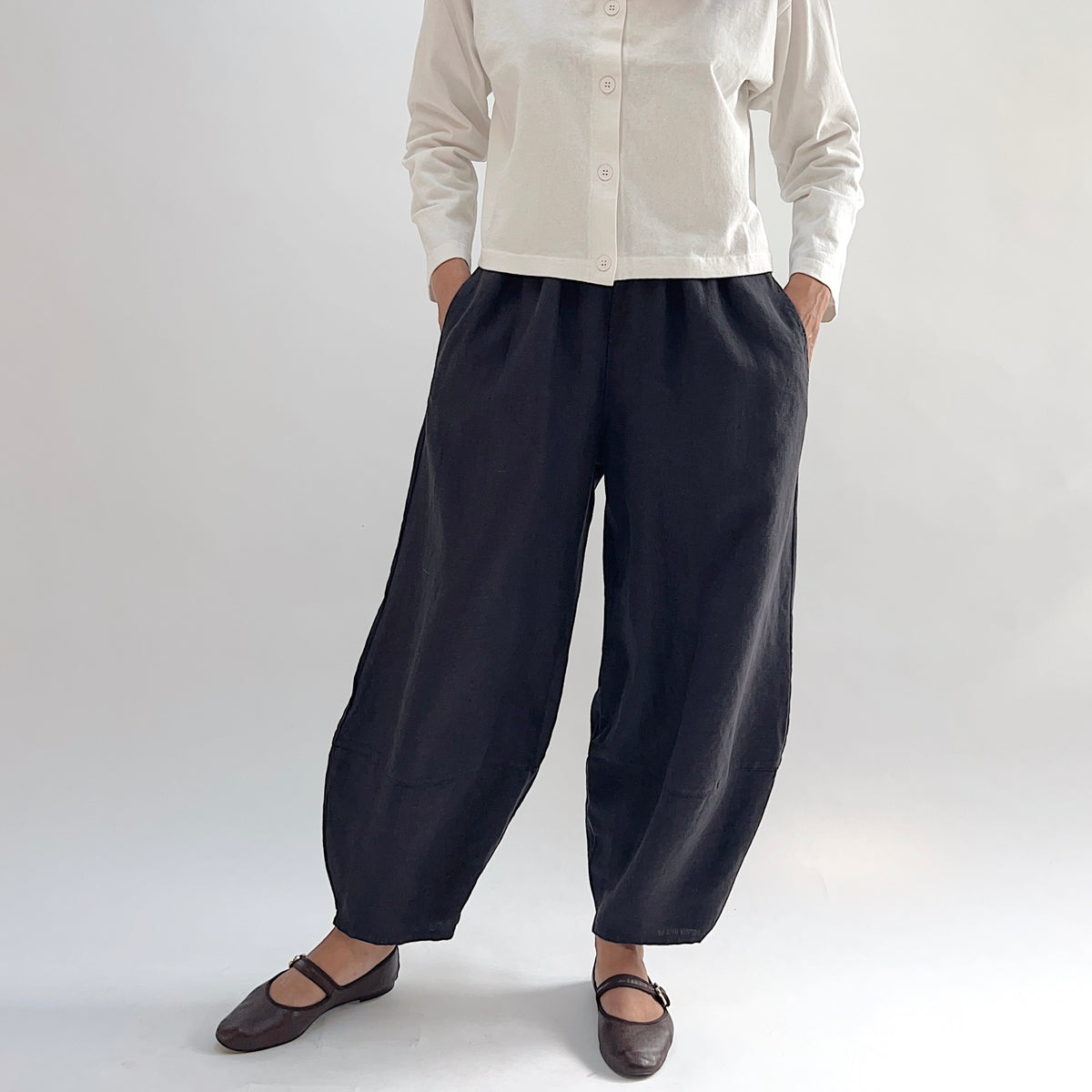 Model with hands in pockets wearing the Bryn Walker Heavy Linen Oliver Pant in Fig, full front view showing barrel-leg silhouette, elastic waistband, side pockets, tapered ankle, and deep fig heavy linen, styled with cream button-up and brown Mary Jane flats
