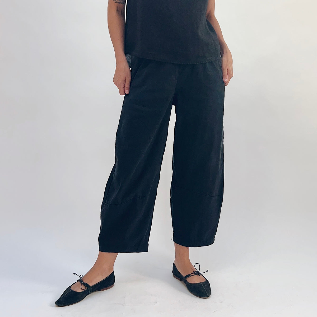 Front view of black linen-tencel lantern pants with wide barrel-leg silhouette, elastic waistband, and side pockets, hands resting in pockets, styled with a black linen top and black bow-tie ballet flats
