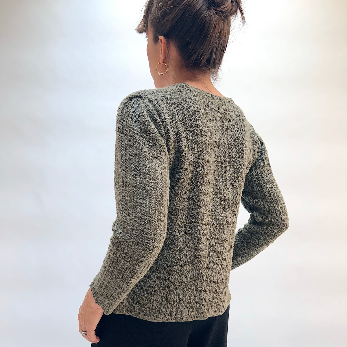 Model wearing the Cut Loose Puff Sleeve Cardigan in Kelp, back view with hair up, showing puffed shoulder seam and textured marled knit of olive green cardigan with long sleeves, styled with black pants
