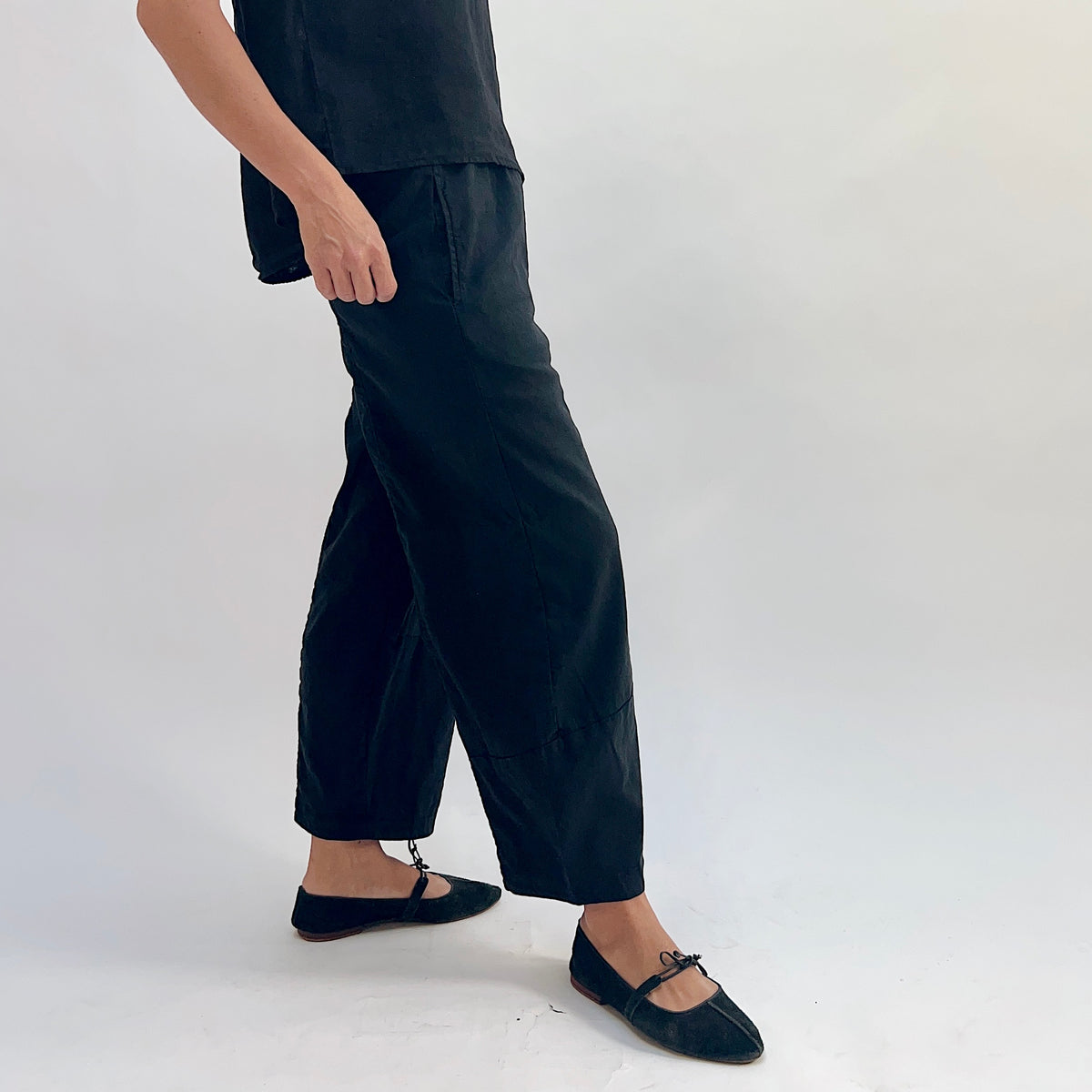 Side view of black linen-tencel lantern pants showing curved seam leg, side pocket, and elastic waistband, worn with a black linen high-low top and black bow-tie ballet flats, hand near hem

