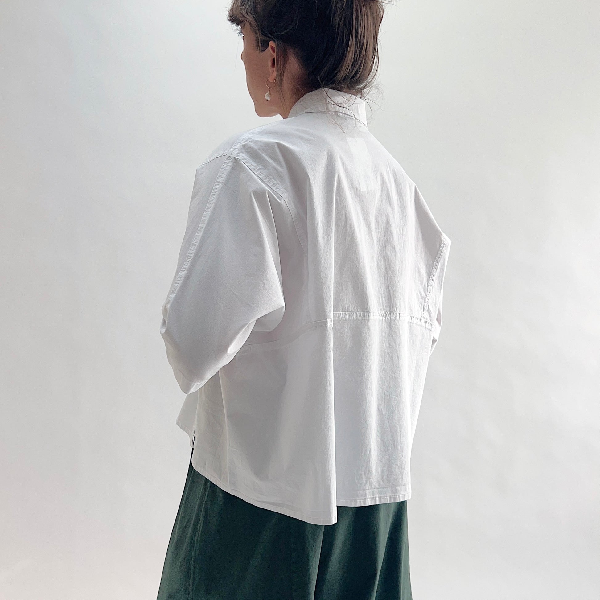 Back view of model wearing Eleven Stitch chalk camp shirt, showing rounded collar, back yoke seam, short sleeves with rolled cuffs, slightly cropped boxy silhouette, and white color, styled with forest green wide-leg skirt