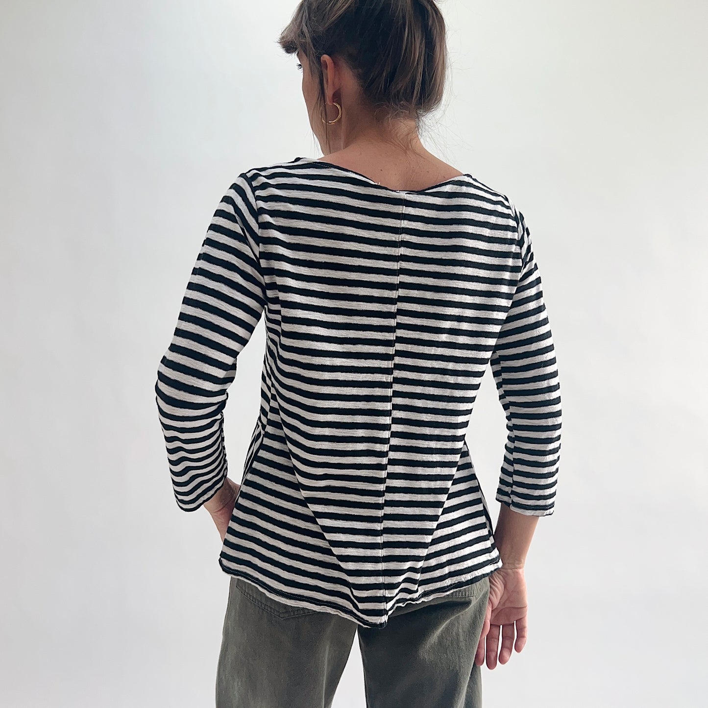 Back view of model wearing a black and white striped A-line boatneck top with 3/4 sleeves, showing the center back seam detail and relaxed A-line hem, styled with olive green pants.
