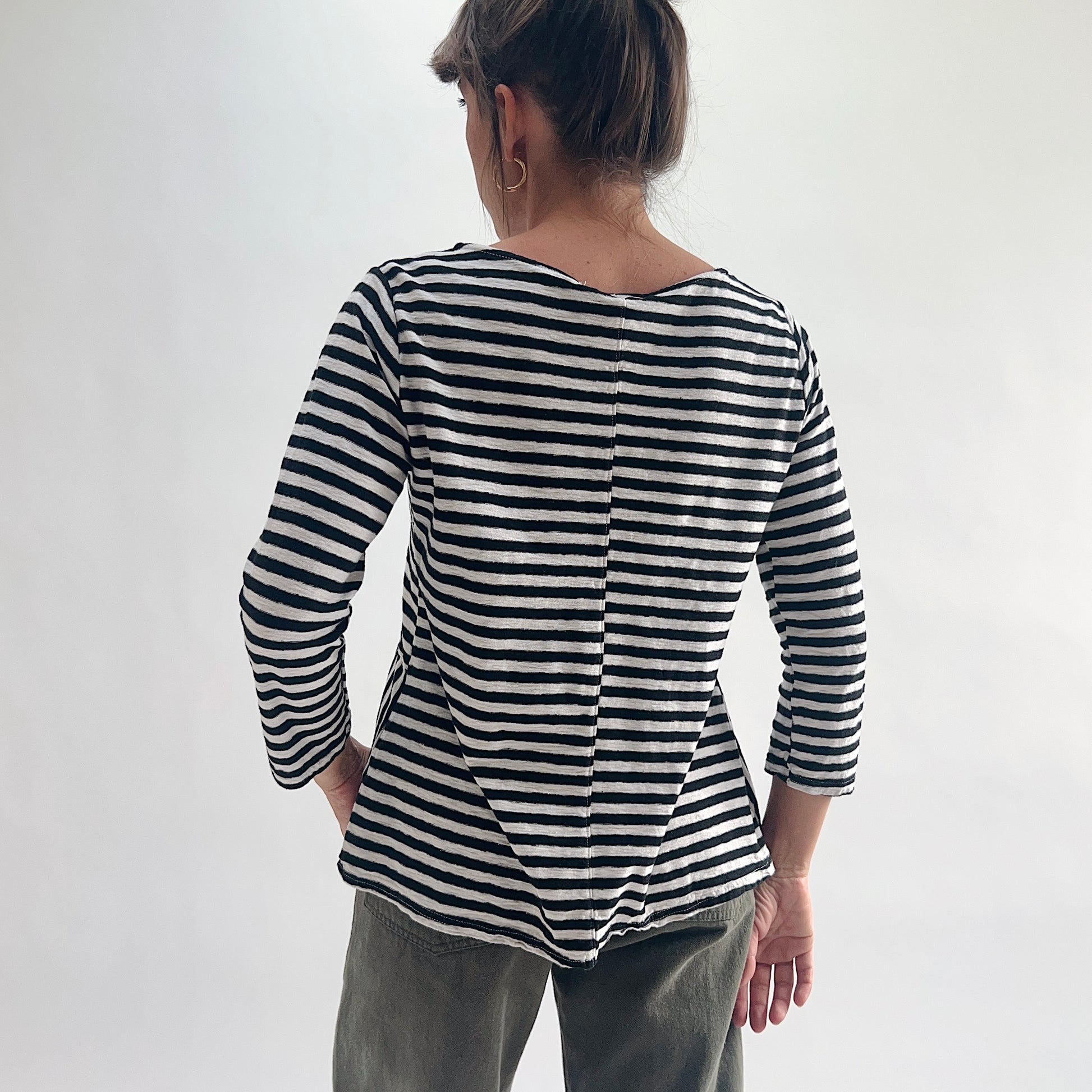 Back view of model wearing a black and white striped A-line boatneck top with 3/4 sleeves, showing the center back seam detail and relaxed A-line hem, styled with olive green pants.

