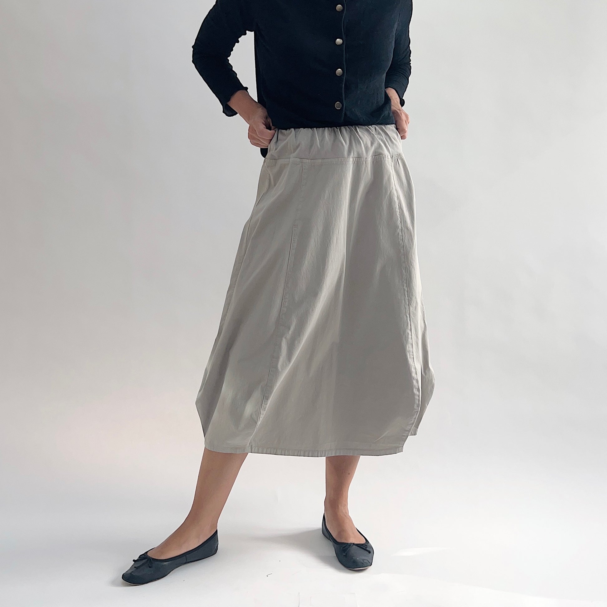 Three-quarter front view of model wearing Eleven Stitch fennel rib yoke skirt with hands on hips, showing elastic knit waistband, curved seam paneling, sculptural bubble hem, below-the-knee length, and warm taupe color, styled with black button-front cardigan and black ballet flats