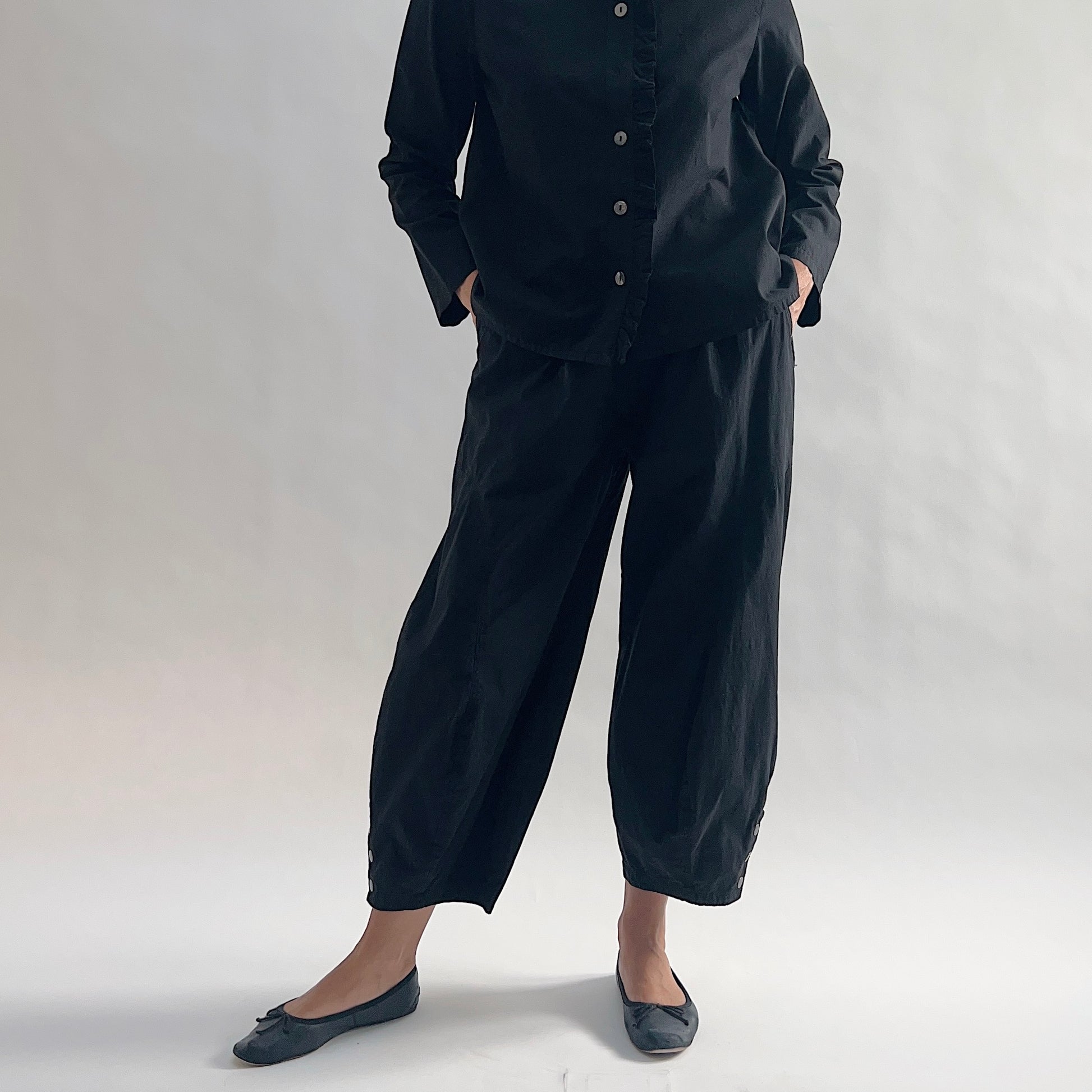Full front view of model wearing Eleven Stitch black snap doreen pant with hands in pockets, showing elastic waistband, wide balloon leg tapering to snap hem at ankle, side seam pockets, and black color, styled with matching black blossom collar shirt and black ballet flats