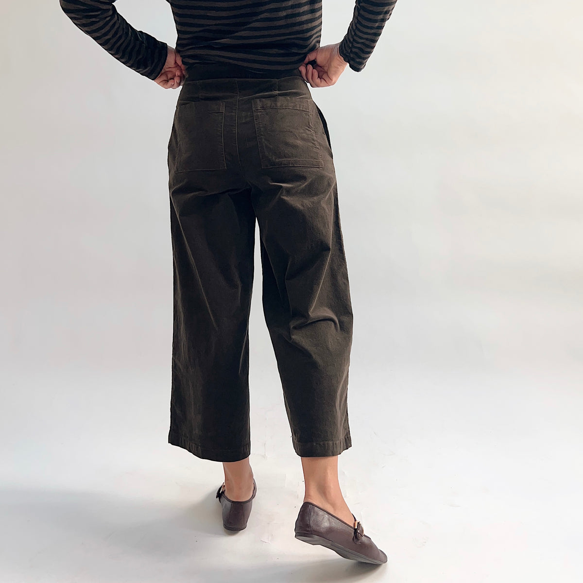 Back view of the Cut Loose Straight Crop Trouser in Chocolate, showing the wide-leg straight crop silhouette, high waist, and patch back pockets in rich dark chocolate cotton-spandex twill, cropped just above the ankle and paired with brown loafers and a striped top
