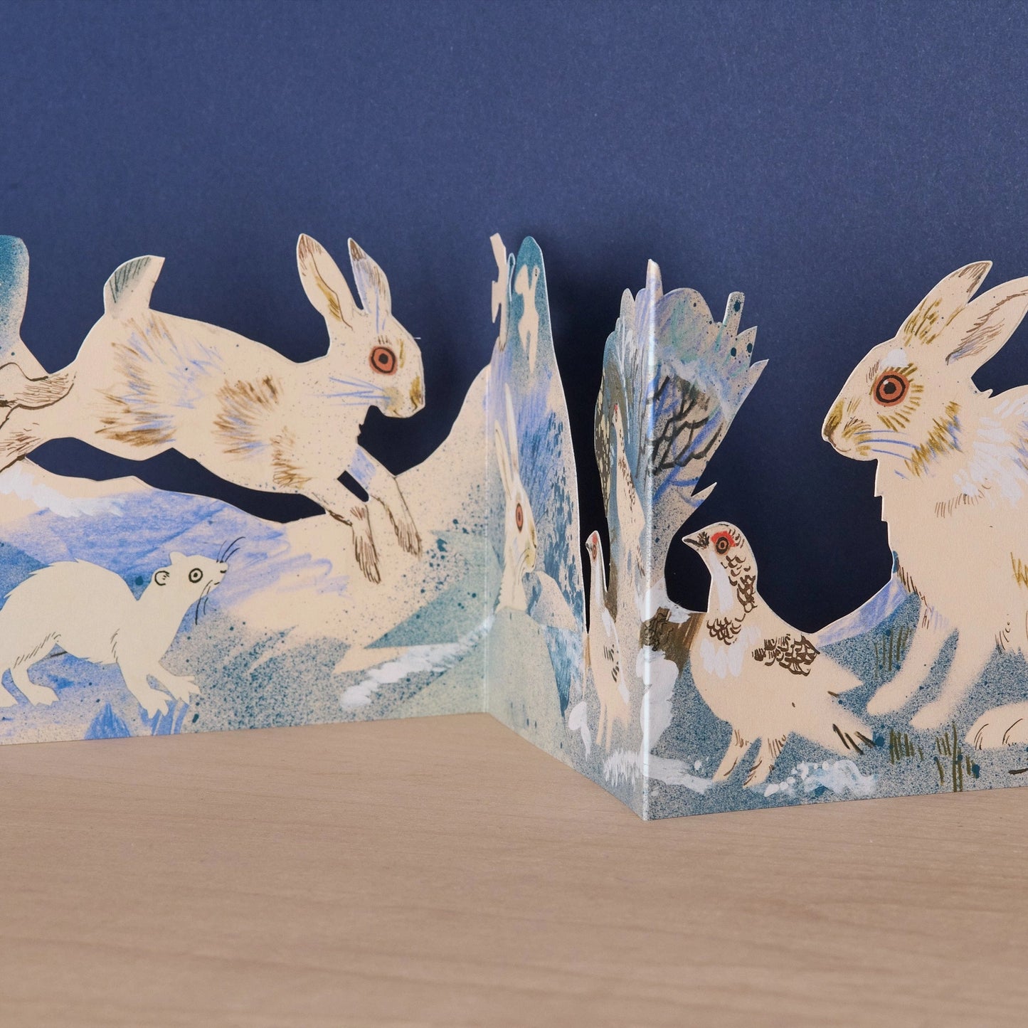 Close-up detail of the Three Fold Winter Wildlife concertina card showing a white hare mid-leap, a stoat, and a ptarmigan bird in a wintry blue and white snowy scene by Mark Hearld