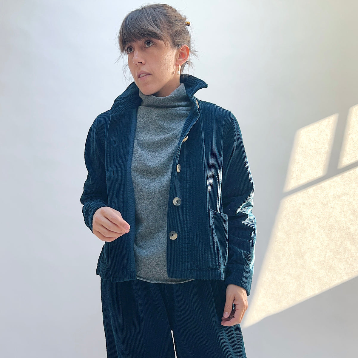 Model wearing the Lux Corduroy Cropped Jacket in Etan open over a gray top, showing the dark teal corduroy and front patch pockets.
