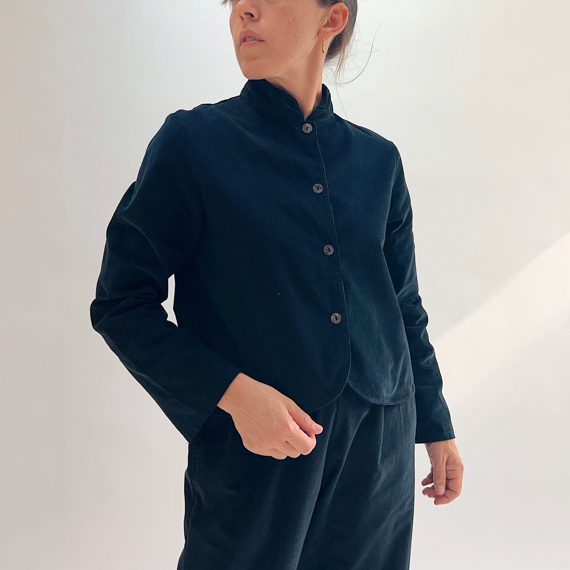 Model wearing the Bryn Walker Corduroy Standing Collar Crop Jacket in Black, angled front view showing ruffled standing collar, shell button closure, wide sleeves, and cropped hem in black pinwheel corduroy, paired with matching black pants
