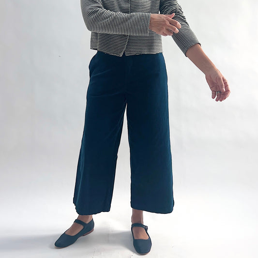 Full front view of model wearing deep teal mini cord straight crop trousers, arms relaxed at sides, showing the flat front, wide straight leg, side pockets, and cropped length, styled with the matching white and black stripe asymmetrical cardigan and teal mary jane flats against a white background.
