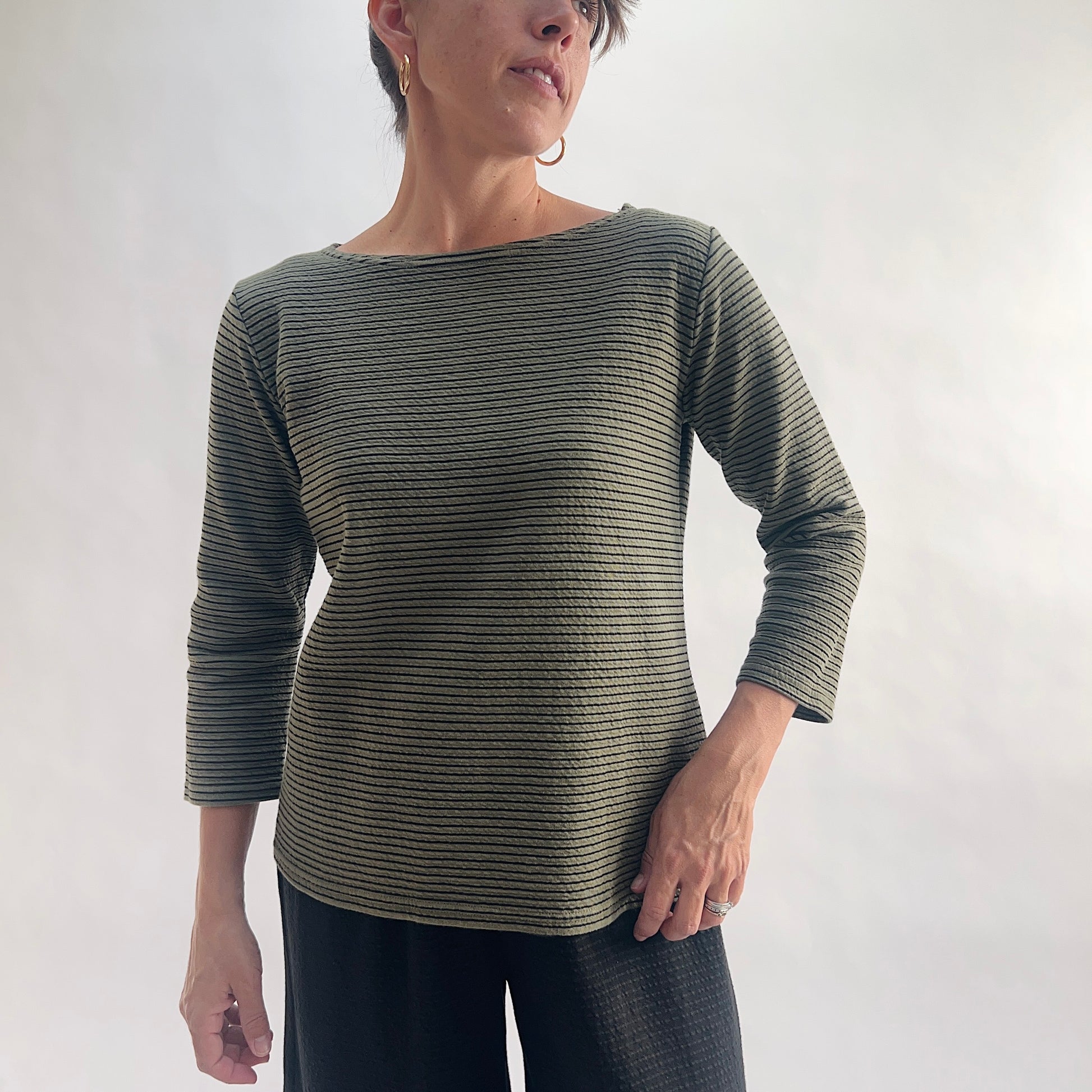 Full front view of model wearing a kelp green tonal textured stripe boatneck tee with 3/4 sleeves, hand on hip, showing the relaxed fit, wide boatneck, and crinkle-texture fabric, styled with black wide-leg pants against a white background.
