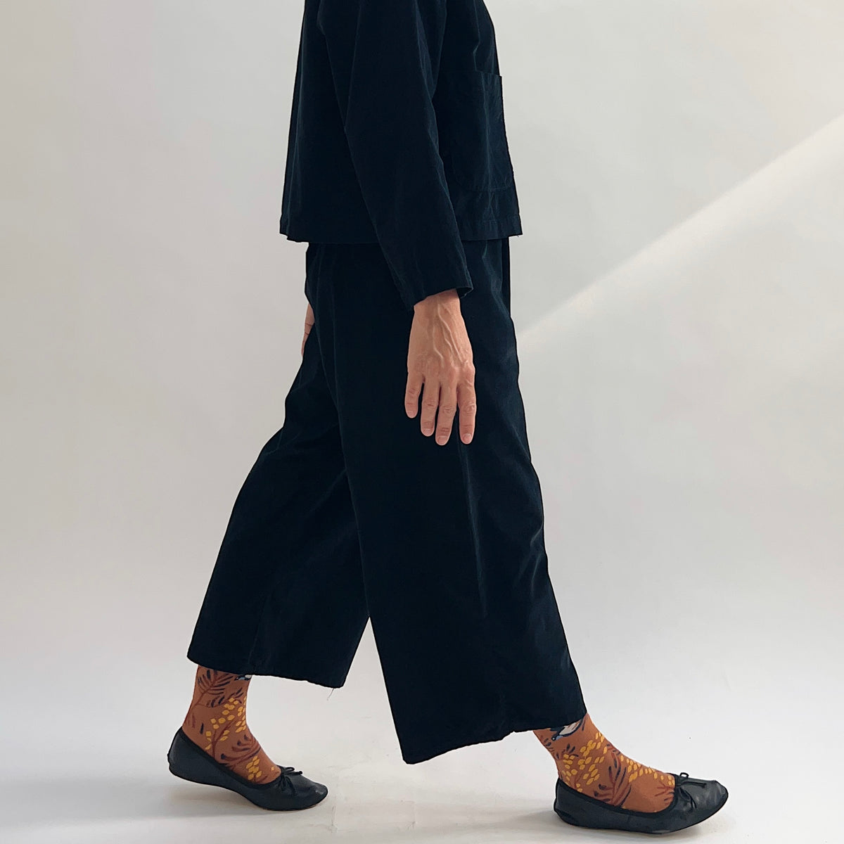 Model walking in the Bryn Walker Corduroy Flood Pants in Black, side profile showing wide-leg silhouette, flood-length cropped hem, and black pinwheel corduroy, paired with matching jacket, printed socks, and black ballet flats
