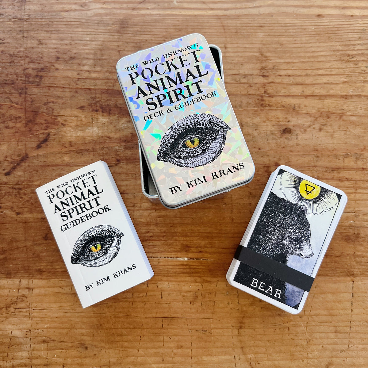 The Wild Unknown Pocket Animal Spirit Deck and Guidebook by Kim Krans, overhead view on a wood surface showing the open holographic tin box, the small white pocket guidebook with reptilian eye cover, and the Bear spirit animal card from the deck
