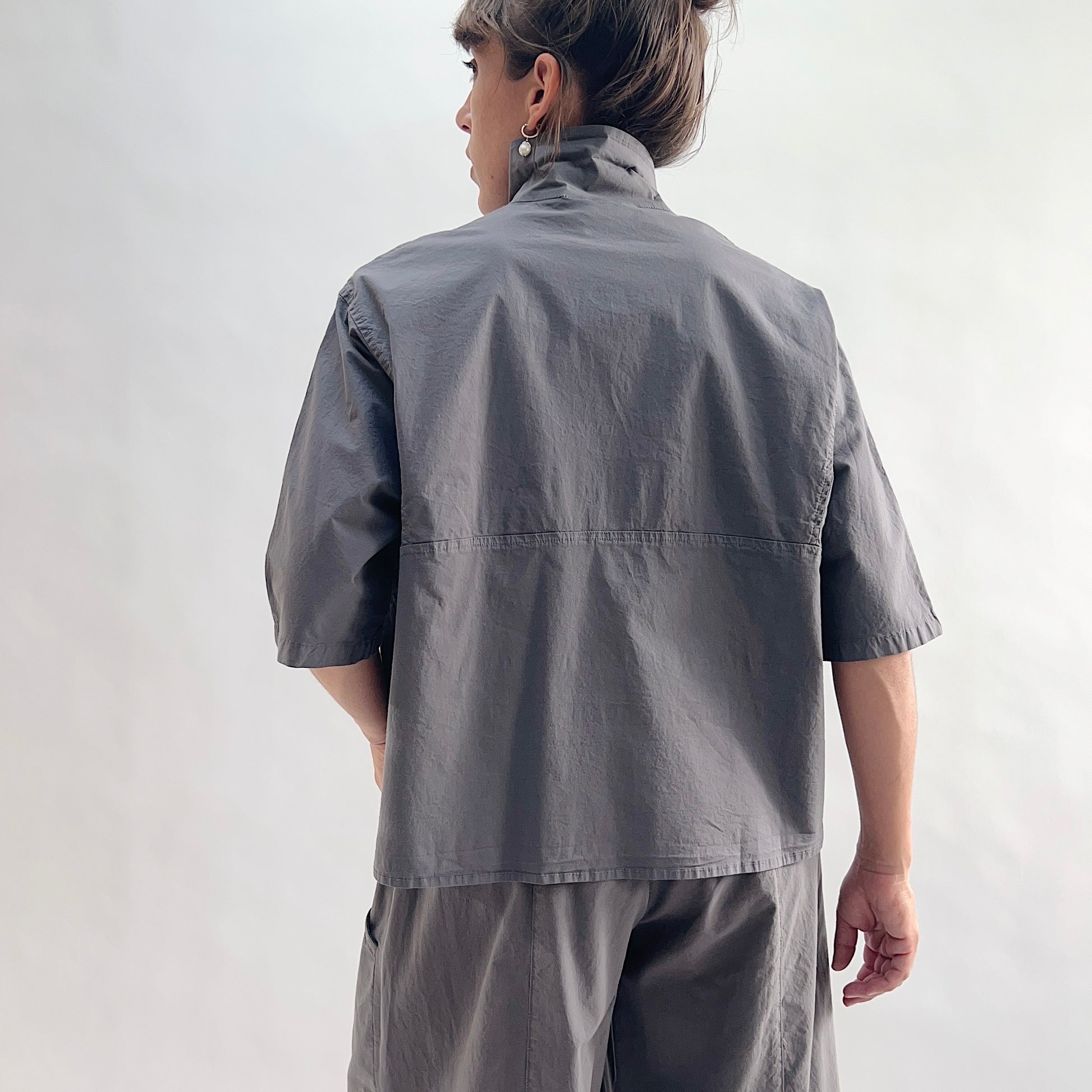 Back view of model wearing Eleven Stitch eiffel tower camp shirt, showing point collar, back yoke seam, short sleeves with rolled cuffs, slightly cropped boxy silhouette, and deep slate grey color, styled with matching eiffel tower one pocket pant