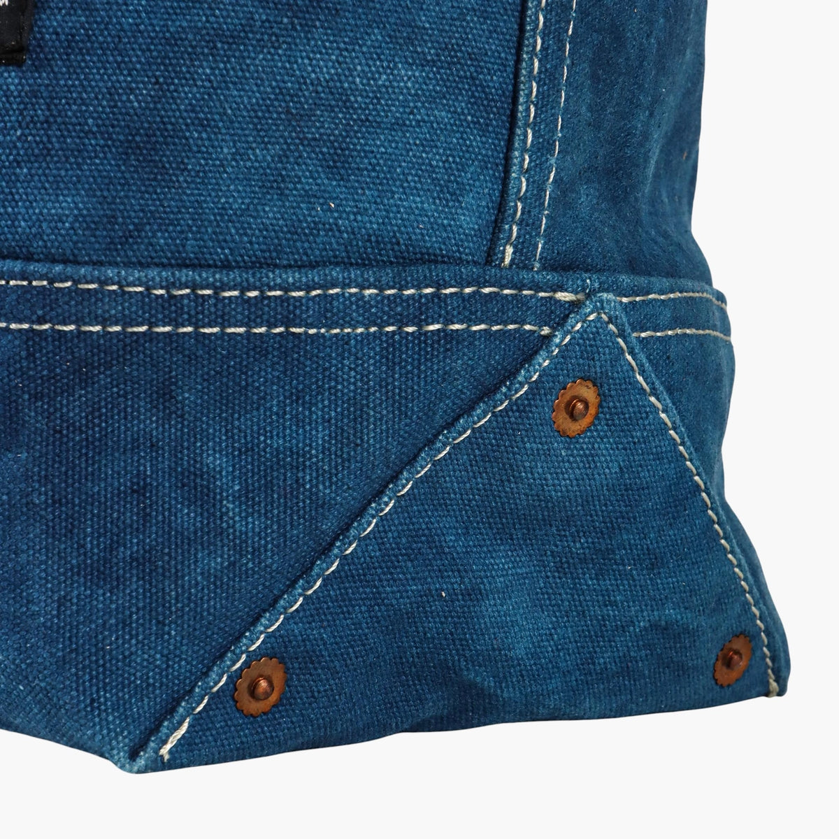 Close-up of the indigo canvas tote's reinforced bottom corner, copper rivets, and double stitching.
