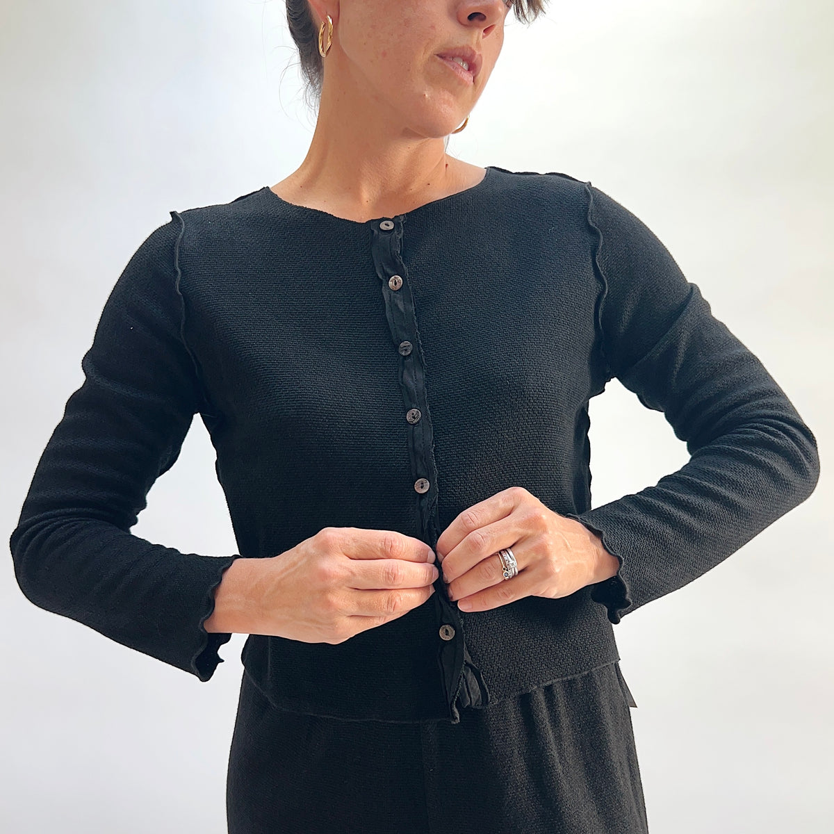 Model wearing the Cut Loose Marled Knit Cropped Cardigan in Black, close-up front view fastening bottom button, showing full button front with dark shell buttons, scalloped lettuce-edge placket, and marled knit texture, styled with a black skirt
