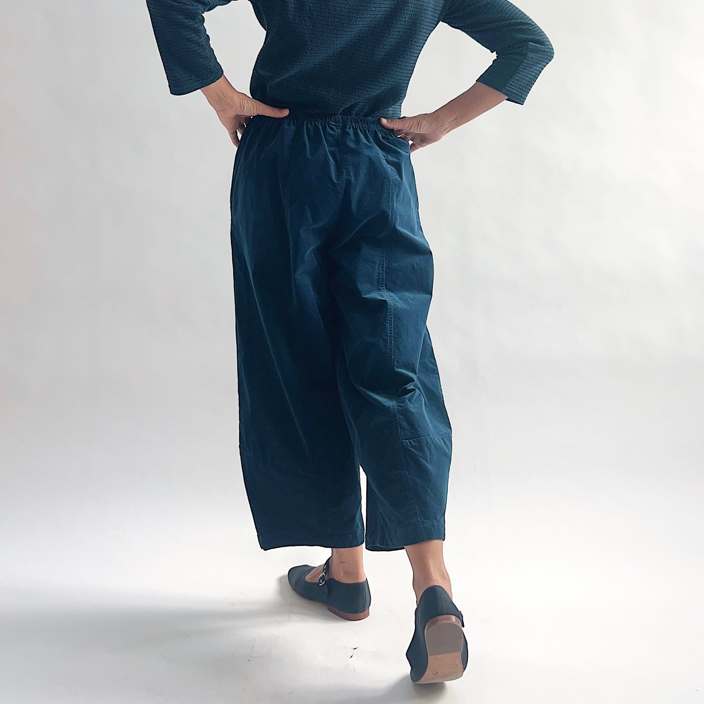 Back view of model wearing deep teal mini cord lantern pants, hands on hips, showing the elastic waist gathering, wide rounded silhouette, side pocket, and tapered ankle, styled with a matching teal textured long-sleeve top and teal mary jane flats against a white background.
