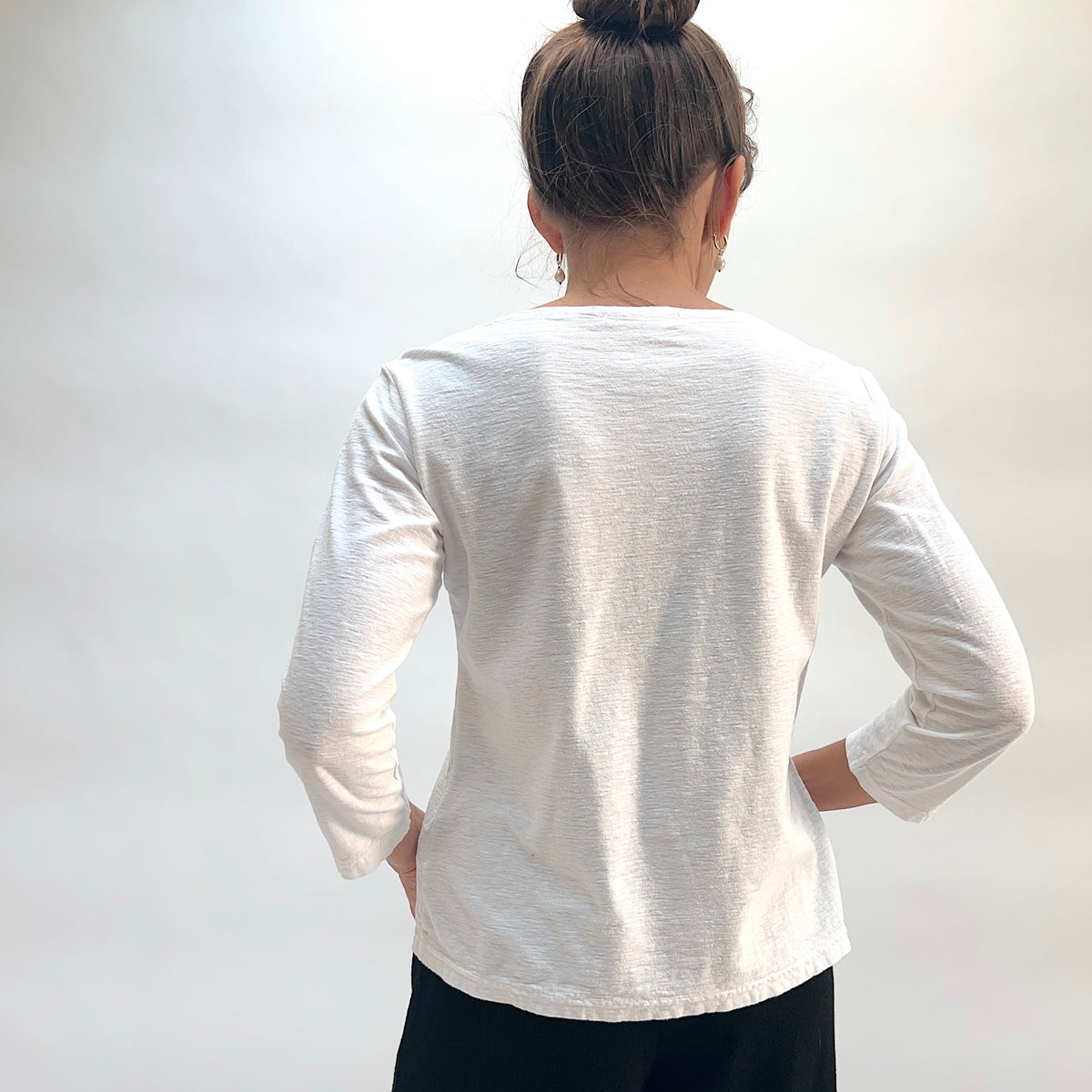 Model wearing the Cut Loose 3/4 Sleeve Boatneck Top in White, back view with hands on hips and hair in bun, showing clean back seaming, side vents, and slub texture of white cotton-linen boatneck top, styled with black pants
