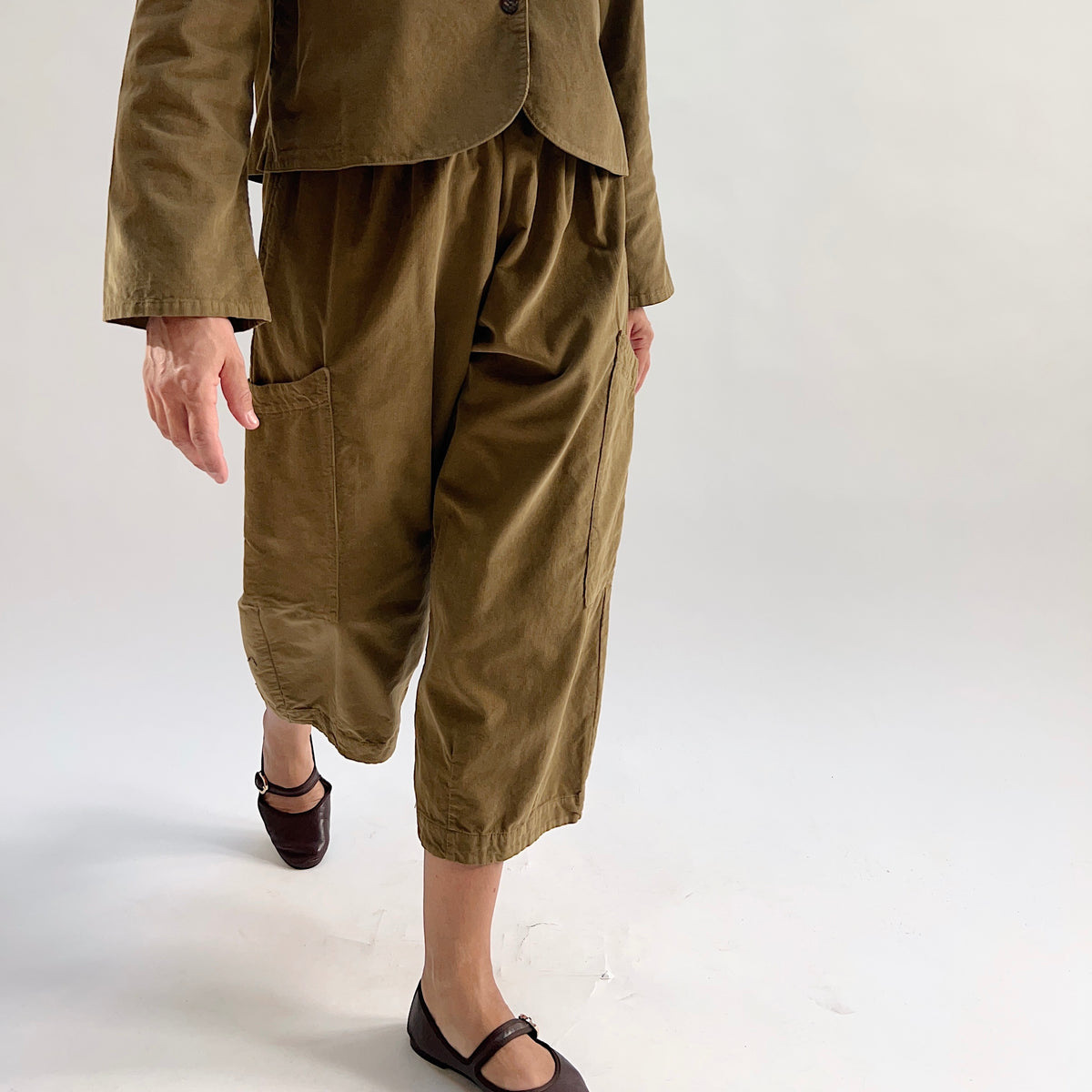 Model in motion wearing the Bryn Walker Corduroy Pasha Pants in Partridge, side view highlighting side leg pocket, cropped wide-leg hem, and olive pinwheel corduroy texture, paired with matching jacket and dark flats
