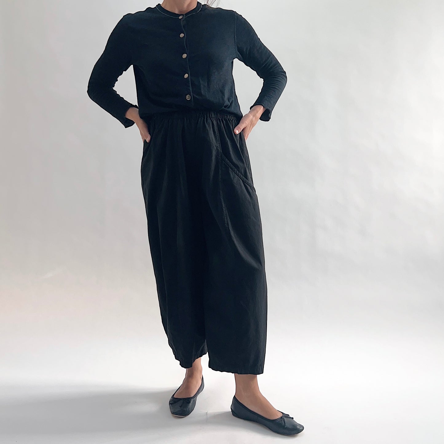 Full front view of model wearing Eleven Stitch black one pocket pant with hands on hips, showing elastic waistband, wide cropped leg, curved side pocket, gathered elastic hem, and black color, styled with black long sleeve button front top and black ballet flats