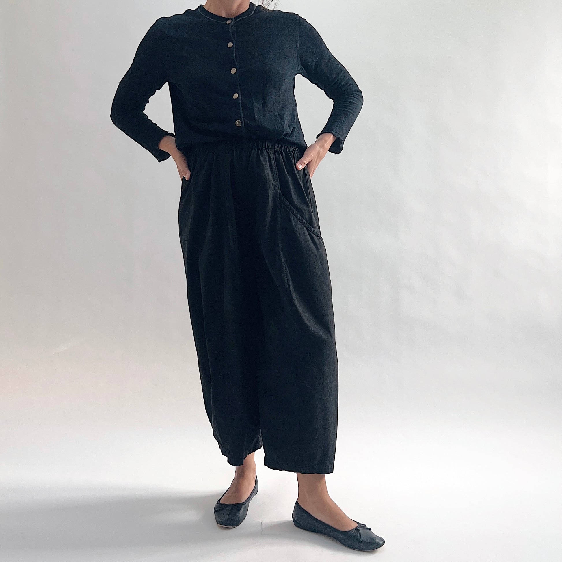 Full front view of model wearing Eleven Stitch black one pocket pant with hands on hips, showing elastic waistband, wide cropped leg, curved side pocket, gathered elastic hem, and black color, styled with black long sleeve button front top and black ballet flats